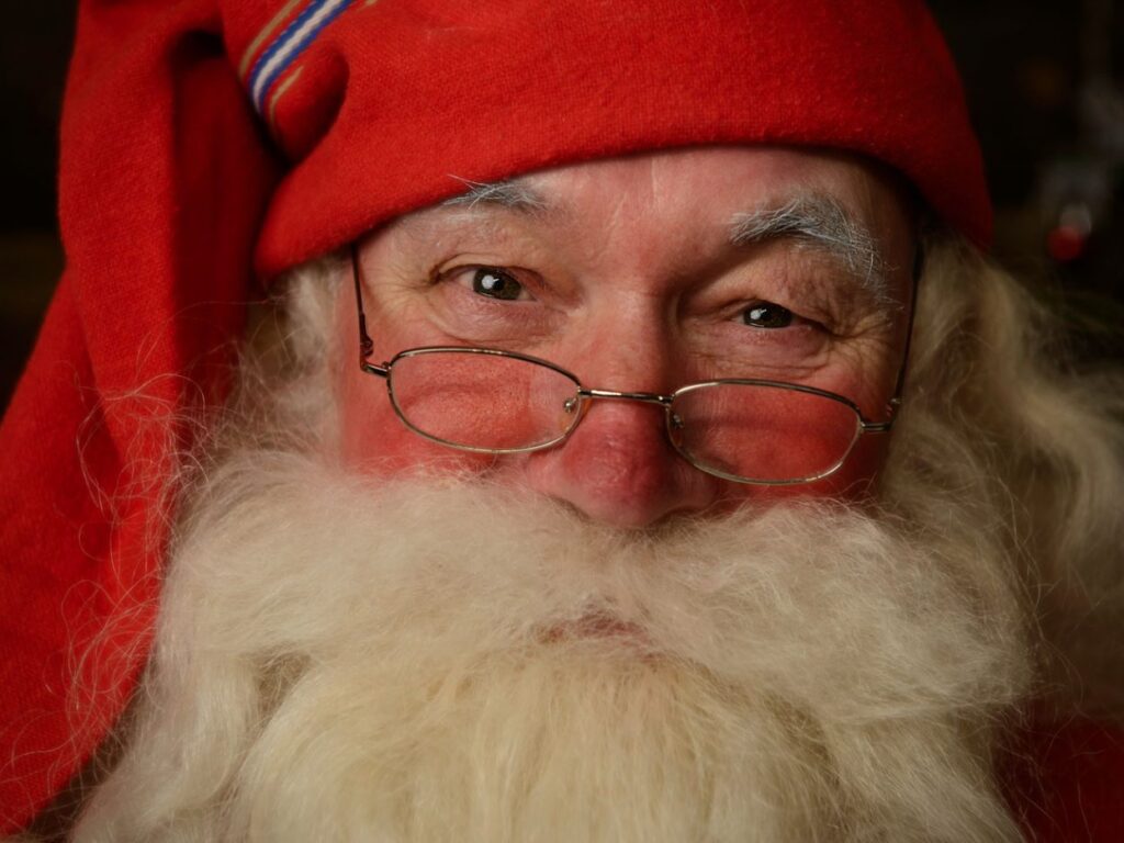 A close up image of Santas smiling face. Santa is wearing a red fabric hat, small gold framed glasses and a full white beard.