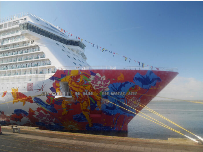Image of the side profile of a cruise ship docked at port. Blue skys in the back ground.
