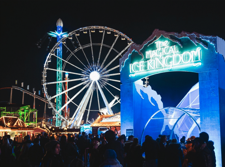 Image of The Magical Ice Kingdom theme park that takes place in Hyde Park London.
A large ferris wheel in the background with an entrance to the right with large neon letters spelling ' The Magic Ice Kingdom'.